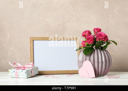 Vaso con le rose rosa, fotogramma vuoto, un dono e un po' di cuori sul tavolo bianco contro luce sfondo marrone Foto Stock