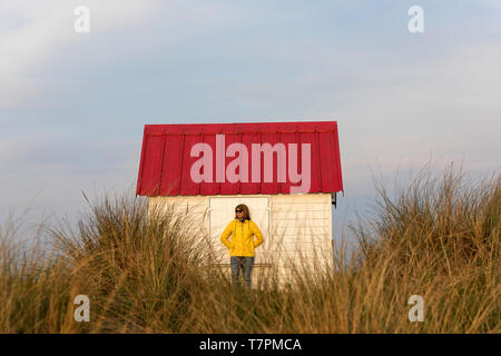 Donna in giacca gialla appoggiata sulla cabina di bianco con un tetto rosso in fila di cabine sulla spiaggia, con tetti colorati a Gouville-sur-Mer, Normandia, Francia Foto Stock