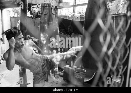 Un kickboxer in Siem Reap Cambogia, mettere in pratica la sua gamba calci contro un sacco di punzone sparato attraverso una catena collegamento recinto e in bianco e nero, mentre gli altri stanno a guardare in background Foto Stock