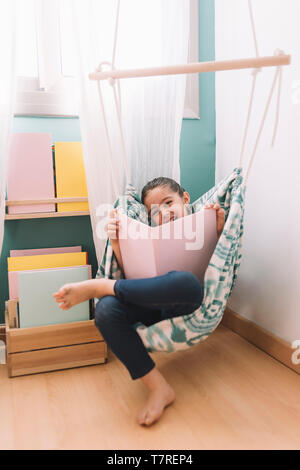 Foto verticale di una piccola ragazza ridere durante la lettura di un libro nella sua camera per bambini vicino alla finestra, funny bel bambino divertirsi a casa, copia spazio per Foto Stock