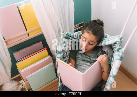 Bambina sorpreso ed entusiasmato la lettura di un libro a casa vicino alla finestra, funny bel bambino divertirsi nella sua camera per bambini, copia spazio per il testo Foto Stock