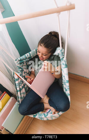 Bambina ride ad alta voce la lettura di un libro a casa vicino alla finestra, funny bel bambino divertirsi nella sua camera per bambini, copiare lo spazio per testo, phot verticale Foto Stock