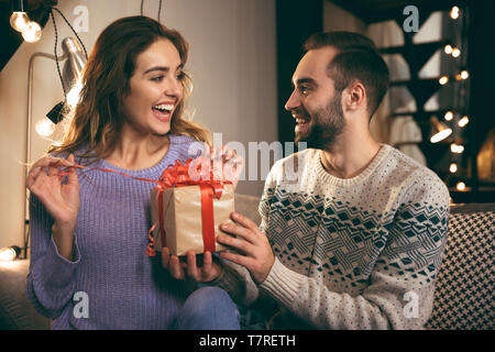 Allegro coppia giovane seduti insieme su un lettino a casa, uomo dando un presente alla sua ragazza Foto Stock