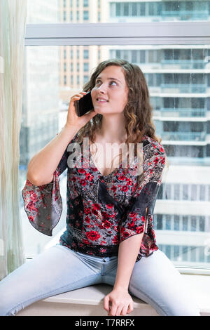 Primo piano della giovane donna felice ragazza seduta in corrispondenza della finestra nella costruzione di parlare al telefono con la sorridente in NYC Foto Stock