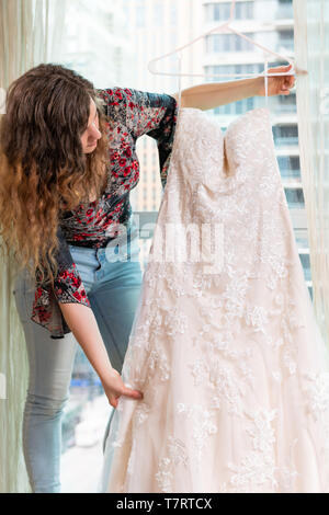 Donna giovane ragazza tenendo guardando abito da sposa su hanger dal davanzale della finestra in ambiente urbano moderno hotel di città alta crescita appartamento condominio prima Foto Stock