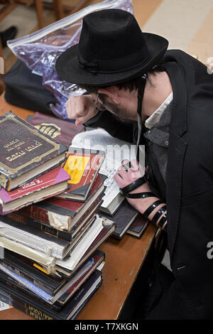 Circondato da una pila di libri, un ebreo ortodosso giovane uomo recita la sua preghiera mattutina mentre indossa i filatteri. A un tempio a Brooklyn, New York. Foto Stock