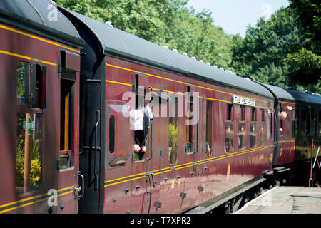 Treno a vapore che arrivano alla stazione. Vista laterale di vintage carrello ferroviario con protezione o ticket inspector sporgersi & guardando fuori della finestra del treno in sun. Foto Stock