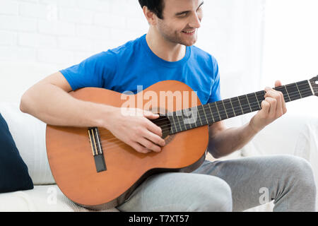 Uomo bello giocare sulla chitarra sul divano di casa. Rilassati guardando uomo seduto su un divano e suonare la chitarra. Foto Stock