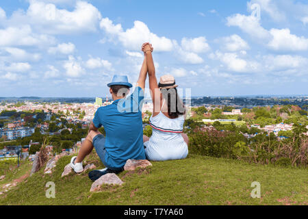 Una coppia di giovani che indossa il cappello con le loro braccia in alto seduta sul pavimento a guardare verso una città da una collina Foto Stock