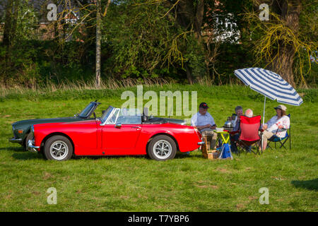 Due coppie anziane fanno un picnic al sole serale accanto alle loro classiche auto sportive inglesi MG & Triumph a Wallingford, Oxfordshire Foto Stock