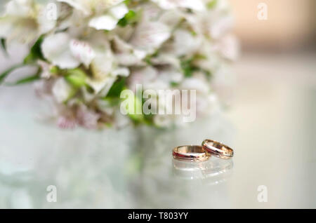 Oro anelli di nozze giacciono su un tavolo di vetro accanto al bouquet Foto Stock
