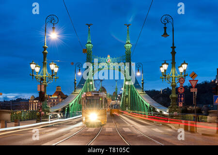 Tram attraversando Ponte della Libertà illuminata di notte con il Gellert Hotel in background, Budapest, Ungheria, Europa Foto Stock