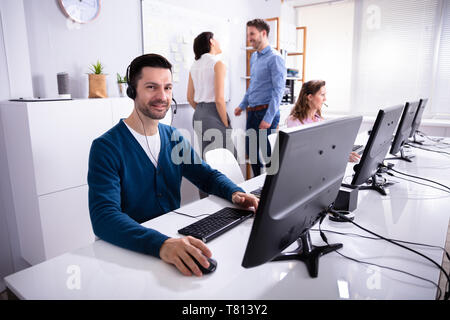Sorridente giovane imprenditore lavora su computer con auricolare in carica Foto Stock