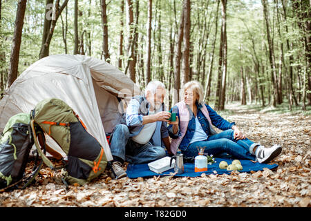 Coppia senior seduti insieme al campeggio con tenda e zaini, godersi la natura e di bere il caffè nella foresta Foto Stock