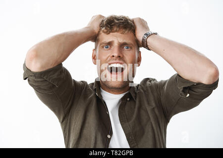 Intensa scioccato guy in stupore con occhi blu e capelli biondi tenendo le mani sulla testa urlando, urlando in preda al panico, freaking out, in piedi storditi e Foto Stock