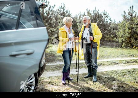 Coppia senior in giallo impermeabili in piedi vicino alla macchina nella foresta, godendo il loro pensionamento Foto Stock