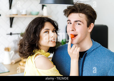 Giovani carino coppia sorridente cucinare insieme alla cucina di casa. Ragazza carina alimenta il suo fidanzato con il rosso dei pomodori di piccole dimensioni Foto Stock