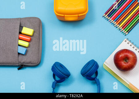 Apple sul quaderno, matite colorate, scatola di pranzo, cuffie, sacchetto custodia a matita con colore pennarelli e un marcatore su sfondo blu. Vista da sopra con copia s Foto Stock