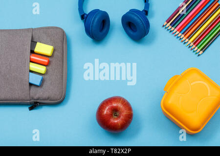 Scatola di pranzo, matite colorate, Apple, cuffie Apple e sacchetto custodia a matita con colore pennarelli e un marcatore su sfondo blu. Vista da sopra con copia spazio. B Foto Stock