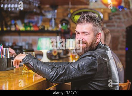 Hipster rilassante presso il bar con birra. Brutale parigamba uomo barbuto sedersi al banco bar bere birra. Ordine di bere alcol. Il bar è un luogo rilassante hanno bere e rilassarsi. Uomo con barba trascorrere il tempo libero nella barra di colore scuro. Foto Stock