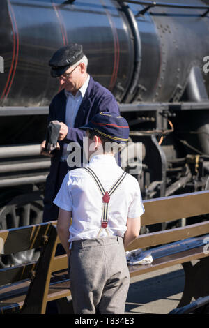 Severn Valley Railway 1940's Wartime Britain. Vista posteriore primo piano, ragazzo della scuola in berretto vintage e stira-pantaloni guardando treni a vapore sulla piattaforma. Foto Stock