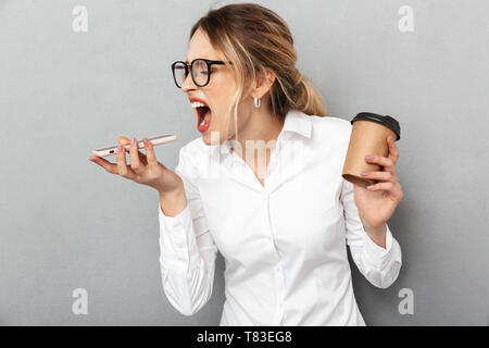 Ritratto di angry imprenditrice che indossano occhiali urlando sul telefono cellulare e tenendo il caffè in ufficio isolate su uno sfondo grigio Foto Stock