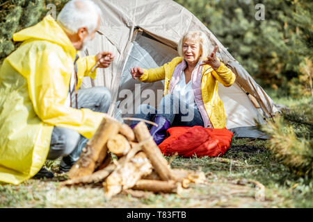 Coppia senior in giallo raincoats divertirsi mentre si effettua il camino presso il campeggio vicino alla tenda nel bosco Foto Stock