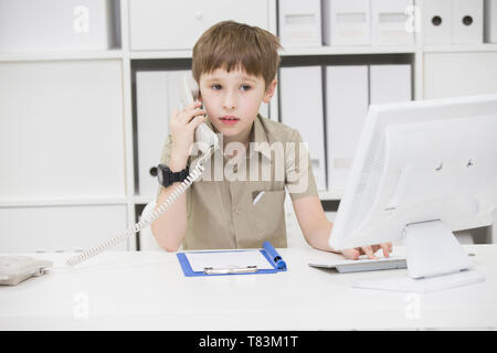 Il bambino al computer tenendo il telefono.Child imprenditore in ufficio.ragazzo giocando imprenditore Foto Stock