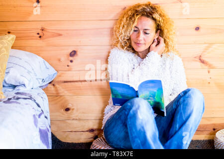 Giovane e bella bionda caucasica curly donna godere la lettura di un libro di carta a casa seduto sul pavimento con pareti in legno in background e letto a Foto Stock