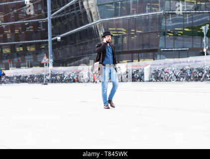 Guidatore di skateboard maschio passeggiando nella piazza della città rendendo chiamata dello smartphone, Freiburg, Baden-Württemberg, Germania Foto Stock