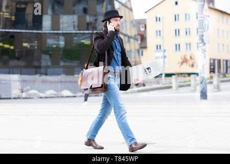 Guidatore di skateboard maschio passeggiando nella piazza della città rendendo chiamata dello smartphone, Freiburg, Baden-Württemberg, Germania Foto Stock