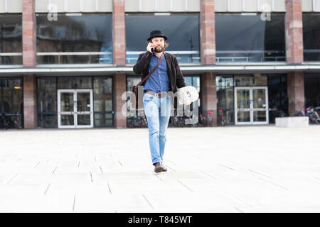 Guidatore di skateboard maschio passeggiando nella piazza della città rendendo chiamata dello smartphone, Freiburg, Baden-Württemberg, Germania Foto Stock