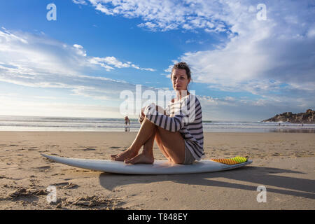 Surfista femmina seduto sulla tavola da surf in spiaggia, ritratto, Cape Town, Western Cape, Sud Africa Foto Stock