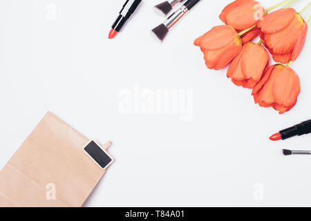 Piana cosmetici laici sfondo della donna rossetti, spazzole e bouquet di fiori accanto alla carta regalo borsa, vista dall'alto sul tavolo bianco. Foto Stock