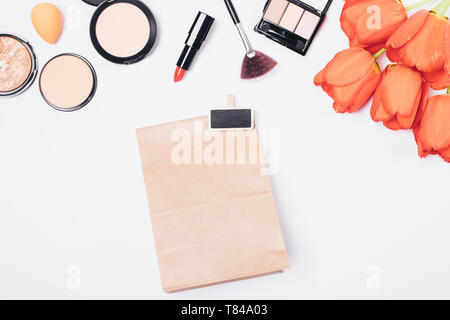 Appartamento laici sfondo per il trucco femminile del rossetto, spazzole e polveri con bouquet di fiori accanto al dono carta marrone borsa, vista dall'alto sul tavolo bianco. Foto Stock