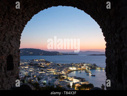 La città di Mykonos, Mykonos, Egeo Meridionale, Grecia. Vista sulla città illuminata e porto attraverso arcata in pietra nel castello dei bastioni, crepuscolo. Foto Stock