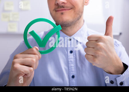 Sorridente giovane imprenditore che mostra pollice in alto segno con icona segno di spunta verde Foto Stock