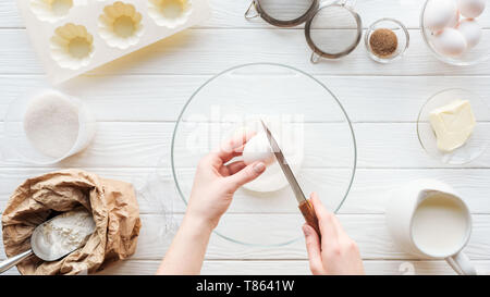 Vista ritagliata della donna con coltello uovo di cracking nel recipiente durante la cottura sul tavolo Foto Stock