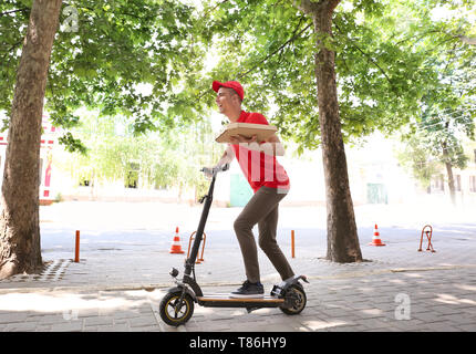 Giovane uomo con Scatola pizza a cavallo di un kick scooter all'esterno. Il cibo del servizio di consegna Foto Stock