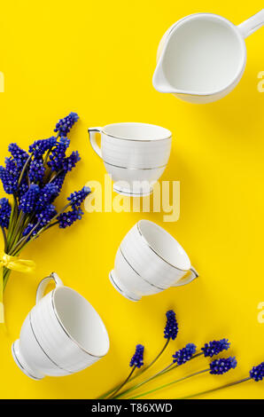 Porcellana Bianca caffè o tè set, jar con latte, muscari fiori su sfondo giallo. Parte superiore verso il basso. Lay piatto. Composizione verticale. Concetto di stoviglie Foto Stock