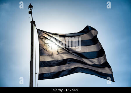 Bandiera nazionale greca Atene retroilluminata // ATENE, Grecia — la bandiera nazionale greca vola contro un cielo luminoso, retroilluminato dal sole ad Atene. Le strisce blu e bianche e la croce della bandiera sono caratterizzate da una silhouette che crea un contrasto sorprendente con la luce del sole dorata. La bandiera, conosciuta come "Galanolefki" (blu e bianco), è il simbolo nazionale ufficiale della Grecia dal 1978 nel suo disegno attuale. Le nove strisce orizzontali rappresentano le sillabe della frase "Eleftheria i Thanatos" (libertà o morte), il motto nazionale durante la guerra d'indipendenza greca. Questo simbolo iconico dell'identità e dell'orgoglio greci Foto Stock