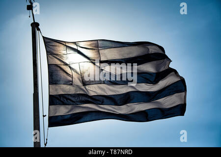 Bandiera nazionale greca Atene retroilluminata // ATENE, Grecia — la bandiera nazionale greca vola contro un cielo luminoso, retroilluminato dal sole ad Atene. Le strisce blu e bianche e la croce della bandiera sono caratterizzate da una silhouette che crea un contrasto sorprendente con la luce del sole dorata. La bandiera, conosciuta come "Galanolefki" (blu e bianco), è il simbolo nazionale ufficiale della Grecia dal 1978 nel suo disegno attuale. Le nove strisce orizzontali rappresentano le sillabe della frase "Eleftheria i Thanatos" (libertà o morte), il motto nazionale durante la guerra d'indipendenza greca. Questo simbolo iconico dell'identità e dell'orgoglio greci Foto Stock