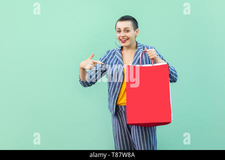 Vuoi? Ritratto di soddisfatto bellissima con capelli corti giovane donna in tuta a strisce in piedi, tenendo shopping bag rossa e puntare il dito su di esso. Foto Stock