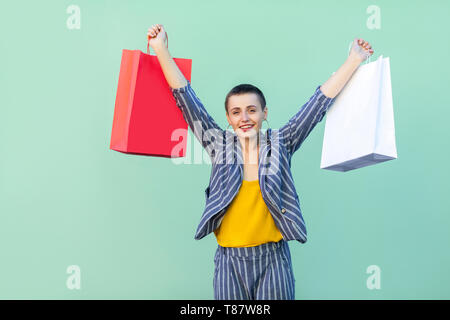 Soddisfatto bellissima con capelli corti giovane donna in tuta a strisce in piedi, tenendo le borse della spesa con i bracci sollevati, sorriso toothy e guardando la fotocamera. Foto Stock