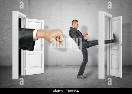 Imprenditore calci porta e grande pugno teso che appare al di fuori della porta aperta sul muro grigio sfondo Foto Stock