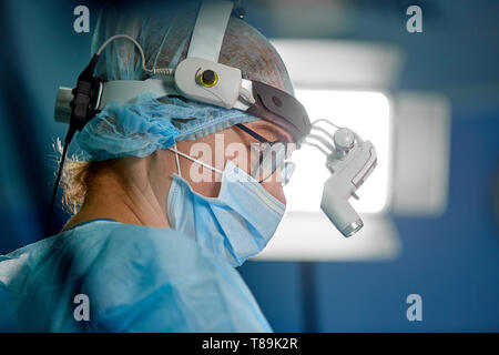 La cura della salute, medico di fondo, chirurgia concetto. ritratto di un chirurgo in con apparecchiature moderne sulla testa durante la chirurgia di close-up, luce di funzionamento. Foto Stock