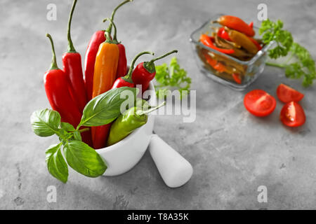 Mortaio con peperoncino fresco i peperoni e le foglie di basilico sul grigio Sfondo testurizzata Foto Stock