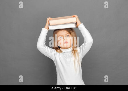 Freestyle. Bambina in piedi isolato su grigio con libri sopra la testa guardando la telecamera in questione Foto Stock