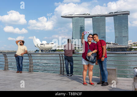 Giovani asiatici carino donna e uomo in stile casual rendendo selfie con fotocamera nella città di Singapore all'esterno. Sullo sfondo il Marina Bay Sands Hotel Foto Stock