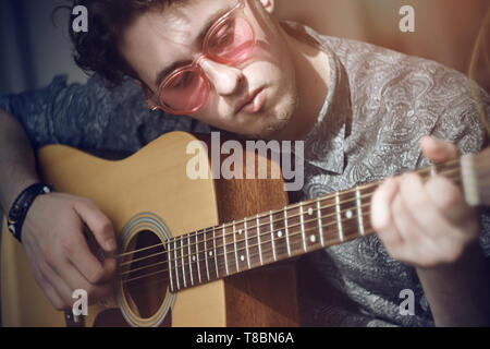 Una parentesi dai capelli ragazzo con la barba e rosa bicchieri, vestito in grigio di una Maglietta modellata, riproduce un di legno chitarra acustica melody, illuminato dalla luce del sole. Foto Stock